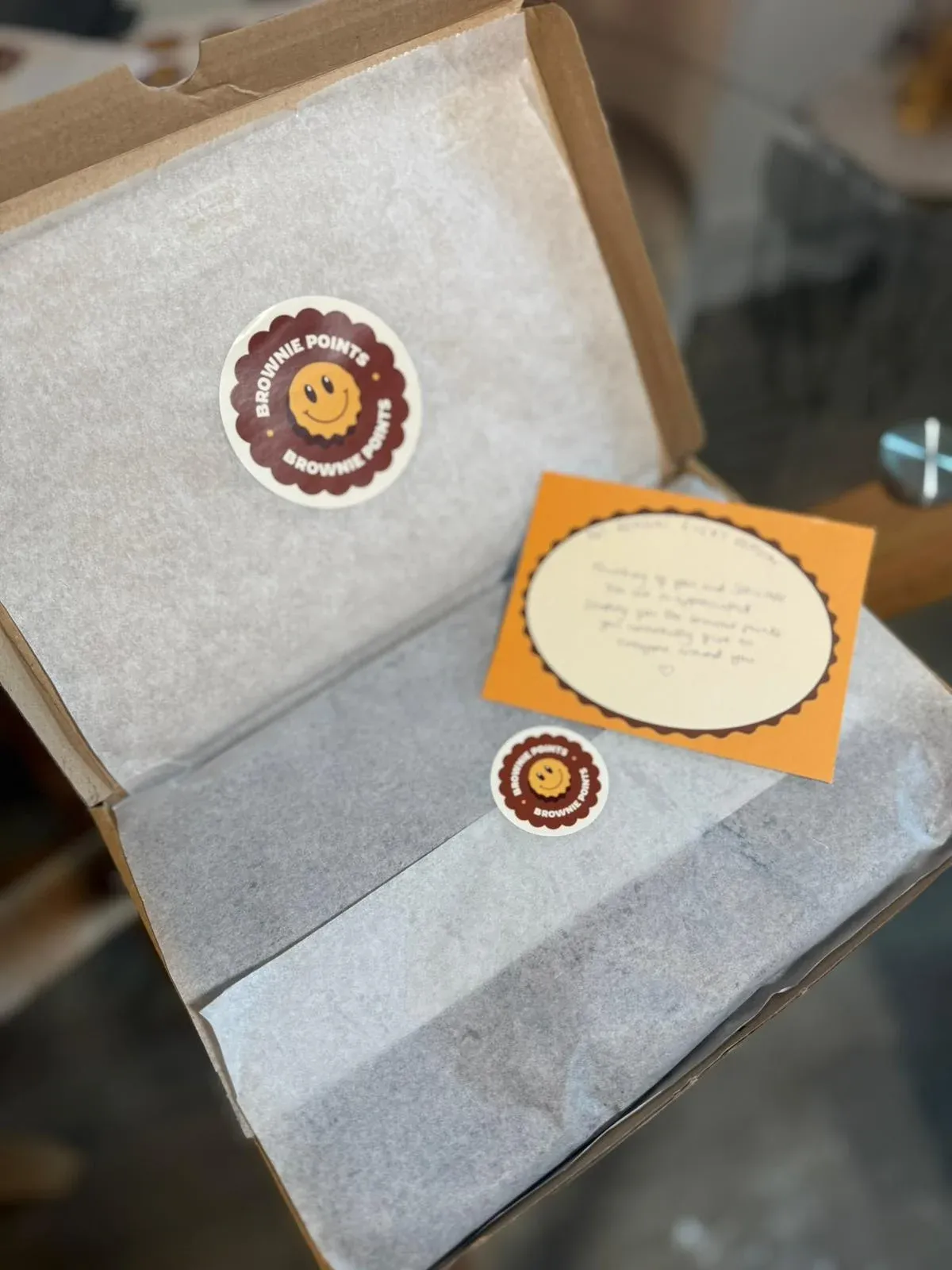 An open Brownie Points box lined with tissue paper, the smiley logo sticker on the lid and a handwritten yellow message card inside.