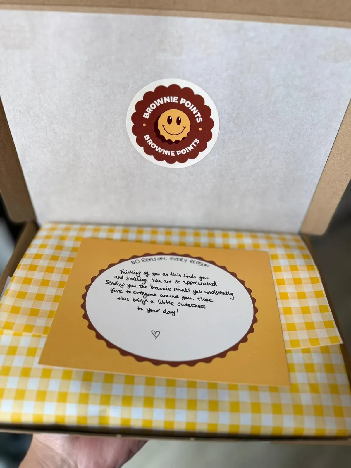 A hand holding an open box: the smiley sticker on the lid, yellow gingham tissue and a handwritten card on top.
